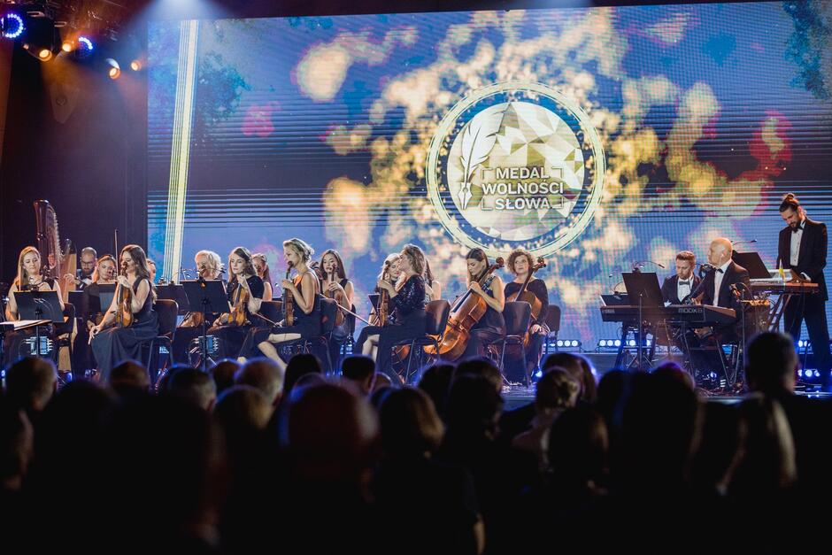 na zdjęciu scena, widać że to uroczysta gala, na scenie siedzi grupa kilkudziesięciu muzyków, w większości trzymają skrzypce