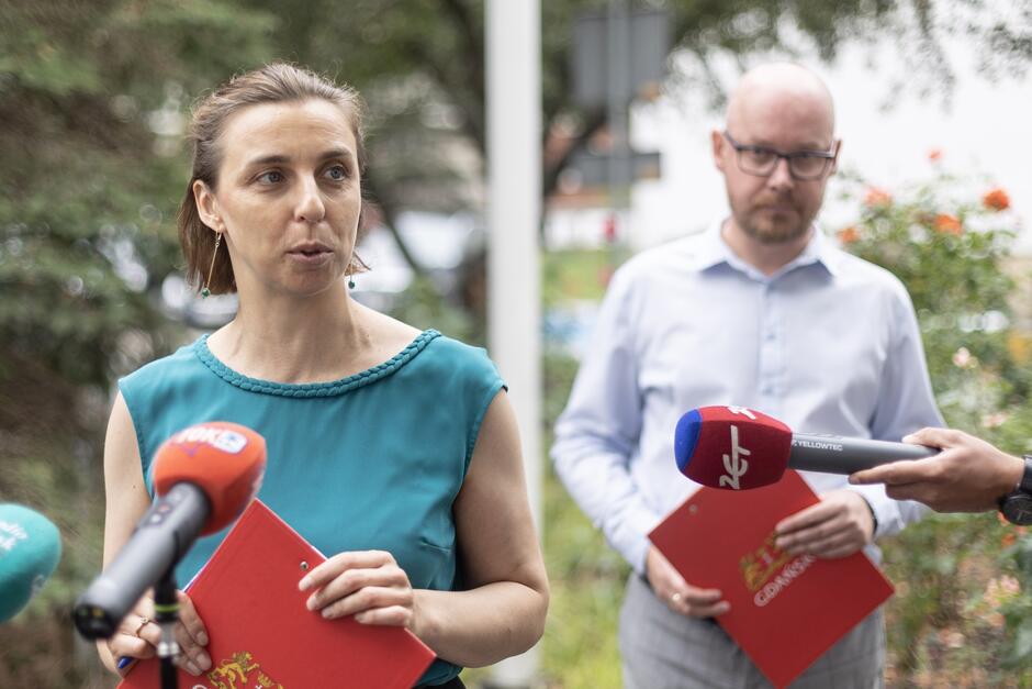 Kobieta występująca na konferencji prasowej, w tle mężczyzna w okularach