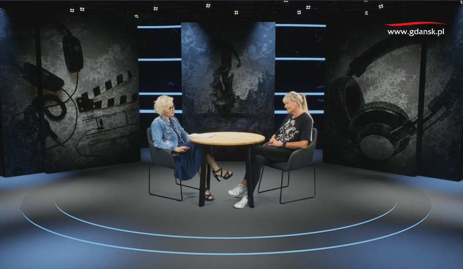 na zdjęciu dwie kobiety w średnim wieku, siedzą na krzesłach przy okrągłym stoliku, w studiu telewizyjnym, rozmawiają