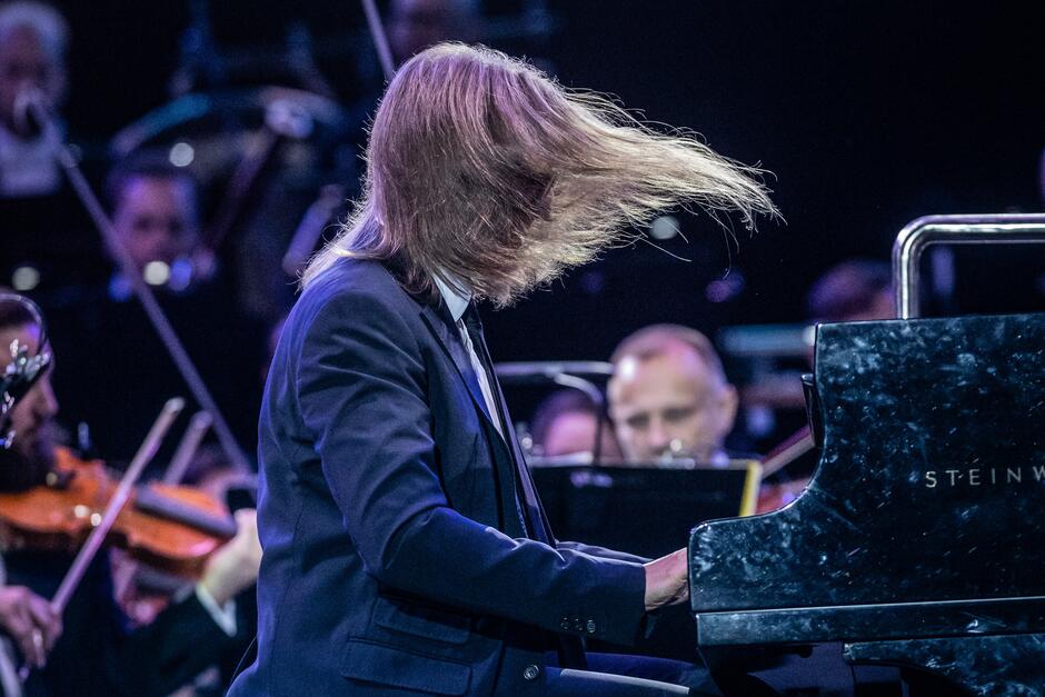 Mężczyzna z długimi włosami i w garniturze siedzi gra na fortepianie