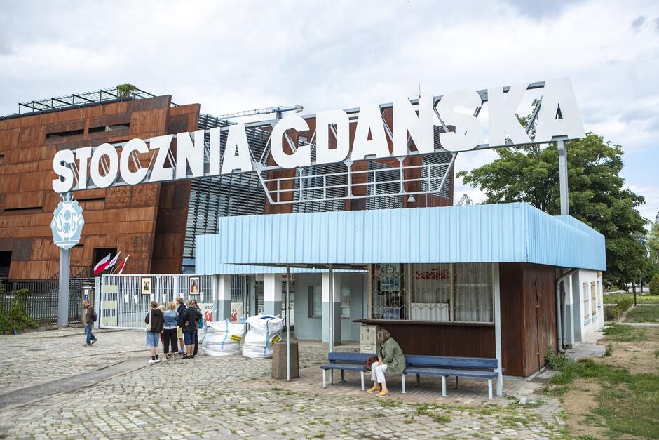 na zdjęciu biały napis stocznia gdańska, znajduje się nad metalową bramą, obok po prawej widać stary kiosk, w tle po lewej fragment rdzawej elewacji gmachu europejskiego centrum solidarności, przed bramą widać grupę kilku stojących osób