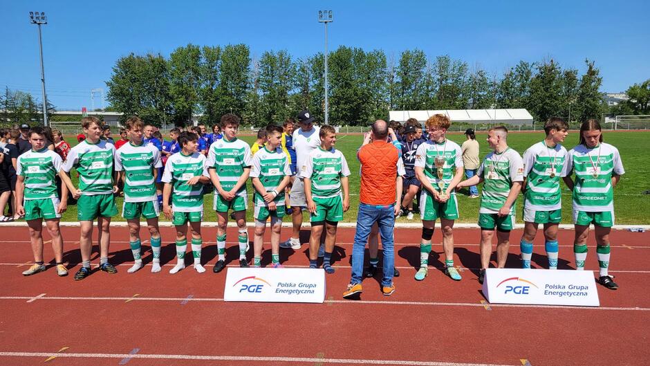 Kilkunastu chłopaków w strojach sportowych stoi obok siebie na bieżni boiska