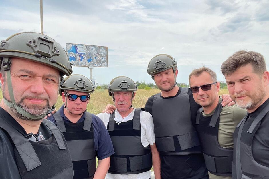 Zdjęcie przedstawia grupę sześciu mężczyzn pozujących do wspólnego selfie. Są w kaskach ochronnych i kamizelkach kuloodpornych 