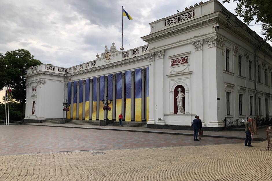 Duży klasycystyczny budynek w jasnym kolorze, w środkowej części jest kolumnada, do której prowadzi kilka stopni schodów. Między kolumnami wisi jedenaście ukraińskich flag, niebieskich w górnej części i żółtych w dolnej 