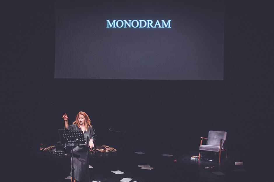 aktorka na pustej scenie, stoi przy pulpicie, z którego czyta, jedną rękę ma uniesioną, w tle wyświetlany napis monodram