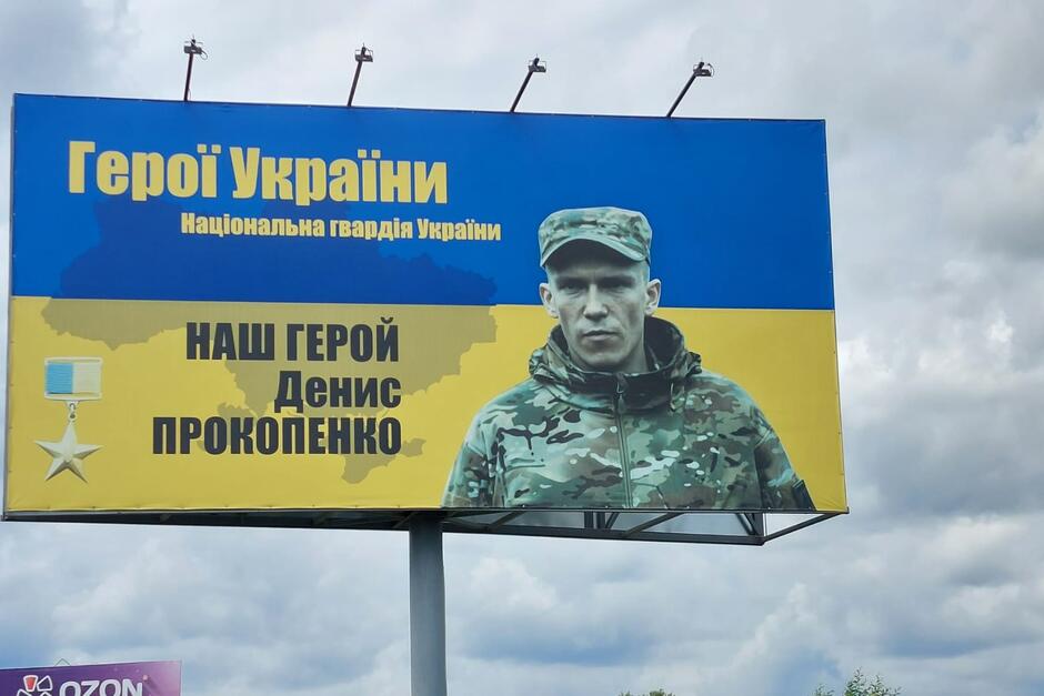 Na billboardzie portret żołnierza w mundurze i czapce, na tle niebiesko-żółtej flagi Ukrainy