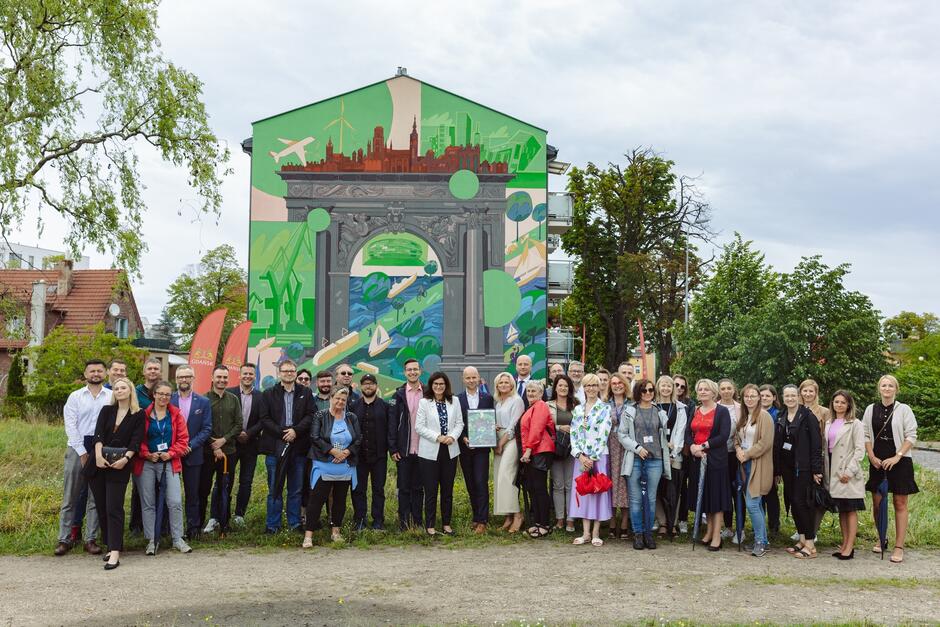 Zdjęcie kilkudziesięciu osób przed muralem na ścianie bloku