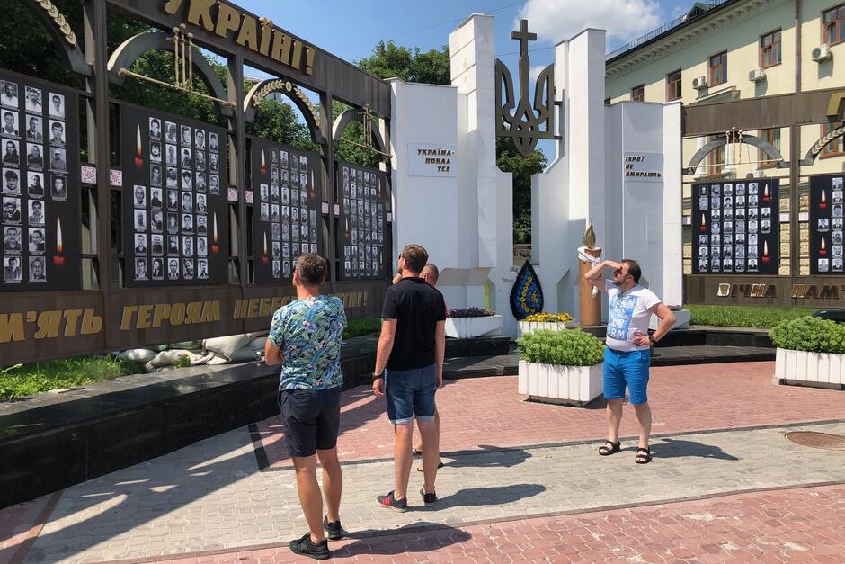 Grupa czterech osób przygląda się skrzydłom mauzoleum, na których umieszczone są fotografie poległych. W środkowej części, między dwoma skrzydłami, widać pomnik w formie godła Ukrainy, które u szczytu zwieńczone jest chrześcijańskim krzyżem 