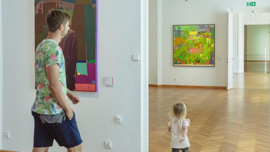 Mężczyzna i mała dziewczynka przemierzają sale galerii. Na ścianach dwa bardzo kolorowe obrazy