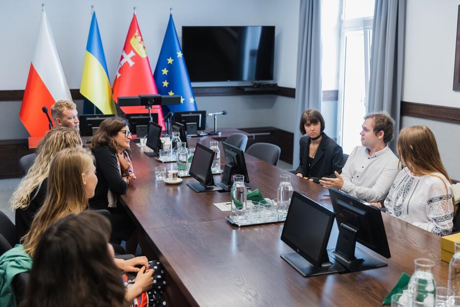 kilka osób siedzi przy długim ciemny stole, po obu stronach, rozmawiają ze sobą, w tle widać cztery flagi, w tym polską, ukraińską Gdańska i Unii Europejskiej