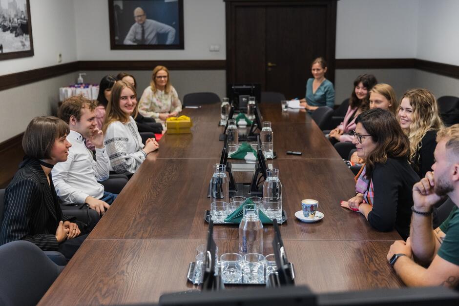 na zdjęciu kilka młodych osób, głównie kobiet siedzi przy długim ciemnym stole, rozmawiają ze sobą