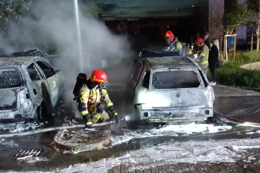 Nocne zdjęcie przedstawia strażaka w trakcie gaszenia dwóch samochodów