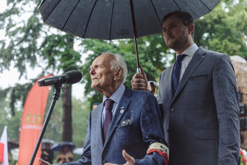 Starszy mężczyzna przemawiający, nad którym parasol trzyma młody mężczyzna 