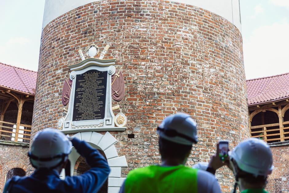 na zdjęciu fragment wieży z widocznym kartuszem umieszczonym nad wejściem, widać też głowy kilkunastu osób stojących przed wieżą