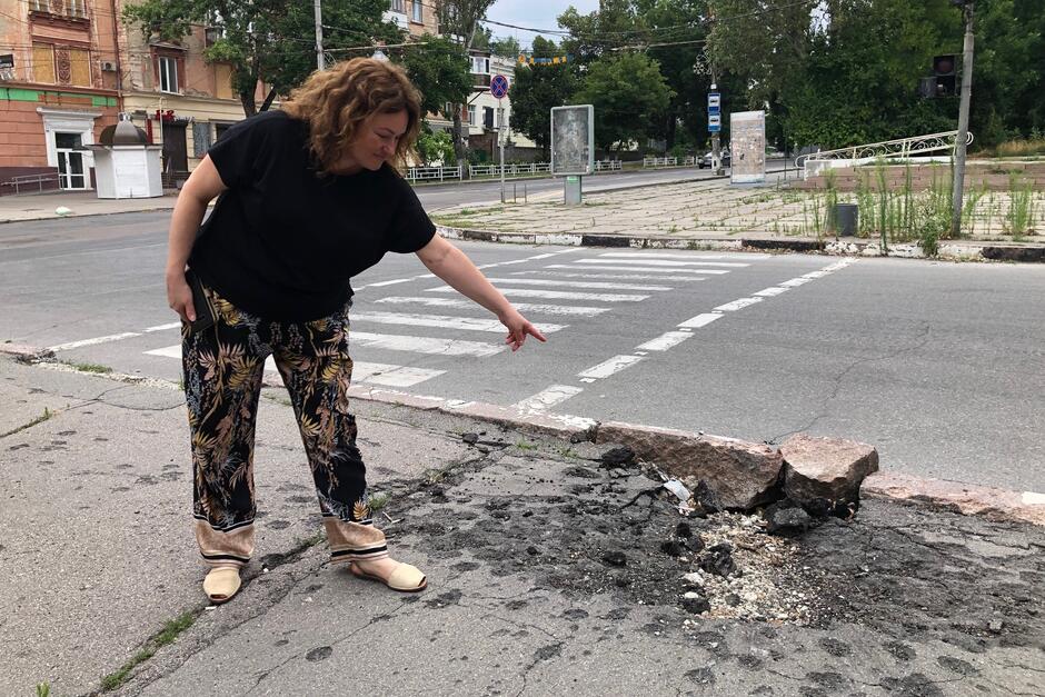 Kobieta stoi na ulicy po lewej stronie, ręką wskazuje na miejsce ze śladami po eksplozji