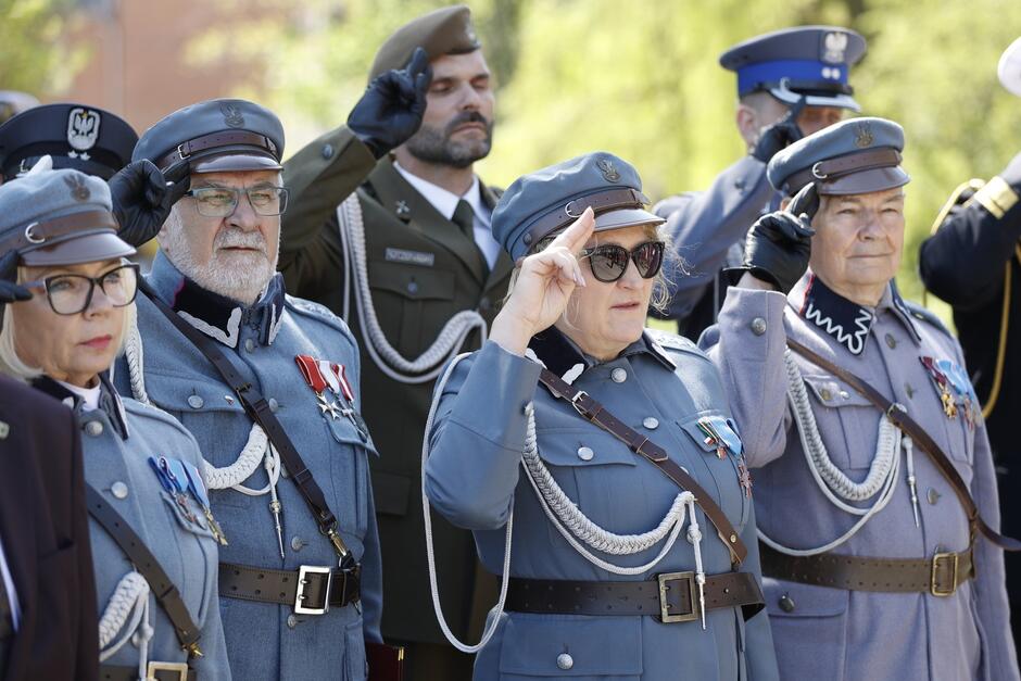 Cztery osoby w szarych piłsudczykowskich mundurach salutują do orzełka na czapkach. Dwie kobiety i dwaj mężczyźni