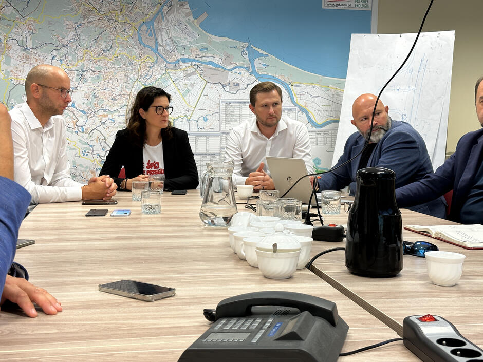 na zdjęciu kobieta i trzech mężczyzn, w marynarkach, wszyscy w średnim wieku, siedzą przy stole, rozmawiają