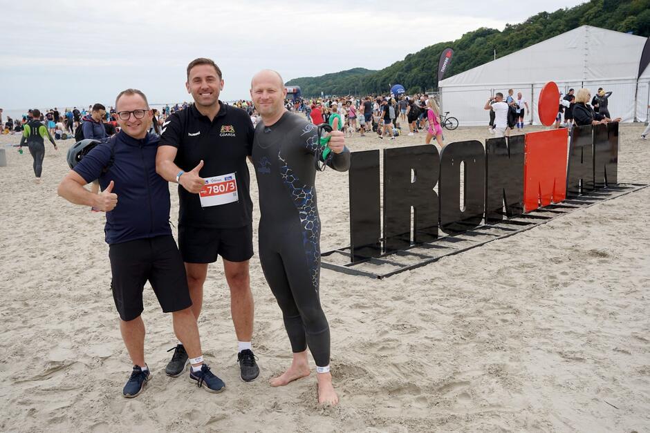 Trzech mężczyzn w strojach sportowych na plaży obok napisu Ironman trzymający kciuki w górze