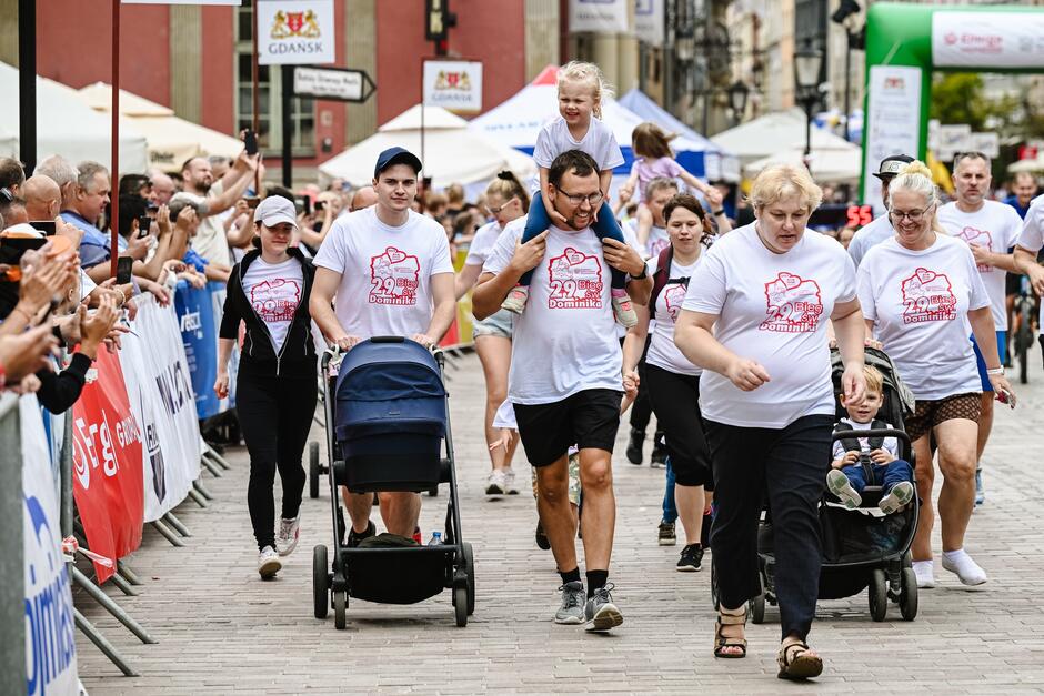 Kilka osób w różnych wieku m.in z wózkiem z dzieckiem biorących udział w biegu na ulicy zabytkowego miasta