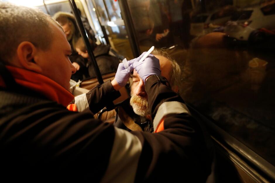 na zdjęciu ratownik medyczny który opatruje twarz bezdomnego mężczyzny