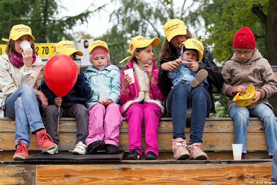 Grupa dzieci w różnym wieku siedzi obok siebie w kolorowych ubrankach i żółtych czapeczkach na głowach