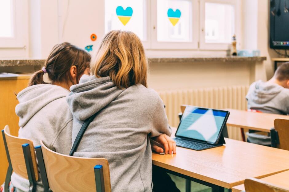 Z lewej strony dwójka dzieci siedzi tyłem i patrzy w ekran laptopa 