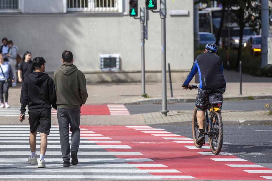 na zdjęciu przejście dla pieszych i obok przejazd rowerowy w kolorze czerwonym, widać jadącego rowerzystę z kaskiem na głowie, obok przechodzi dwoje pieszych