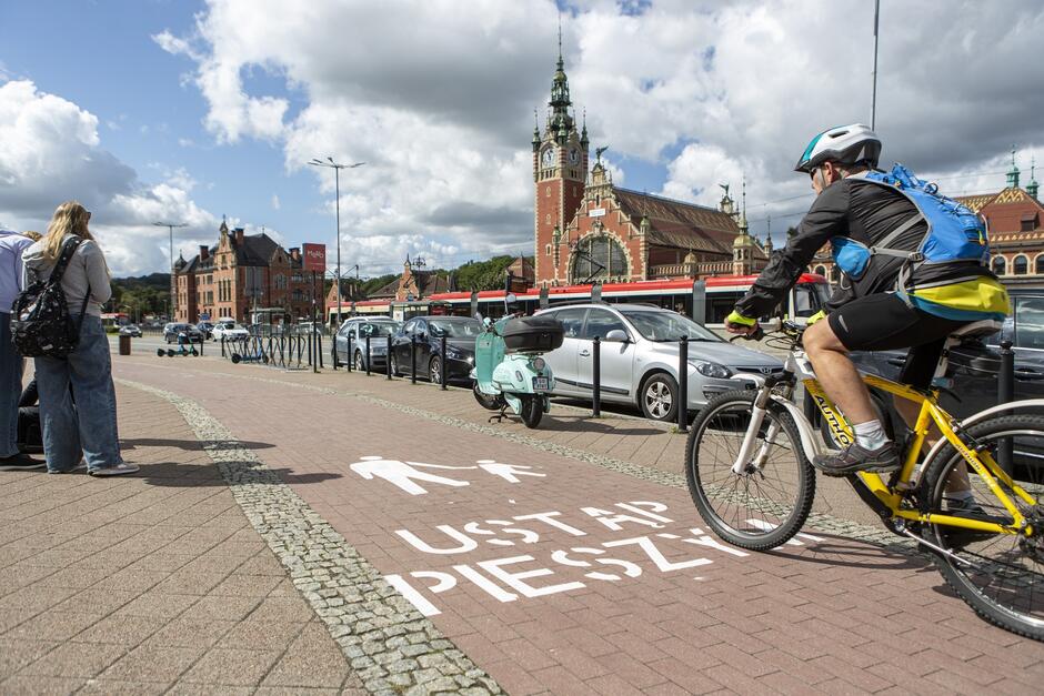 na zdjęciu rowerzysta przejeżdżający drogą rowerową na której jest napis ustąp pieszym, po lewej stoją dwie osoby, w tle po prawej widać budynek dworca