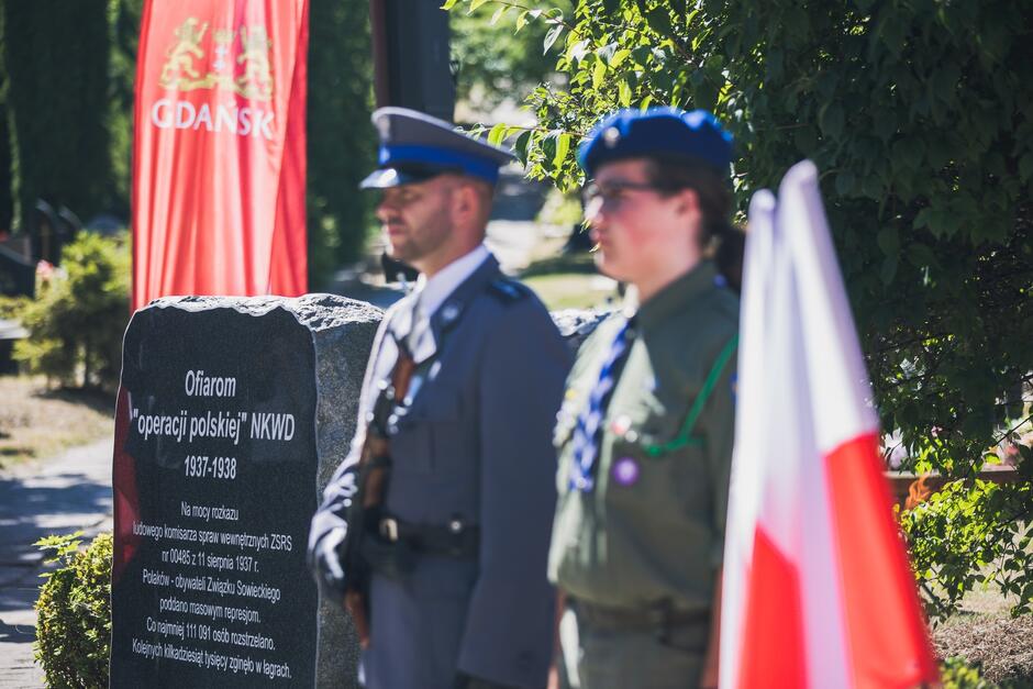 Na warcie stoją dwie osoby w mundurach policjant i harcerka. Na lewo od nich znajduje się czarny marmurowy obelisk z napisami poświęconymi ofiarom akcji polskiej NKWD w latach 1937-1938