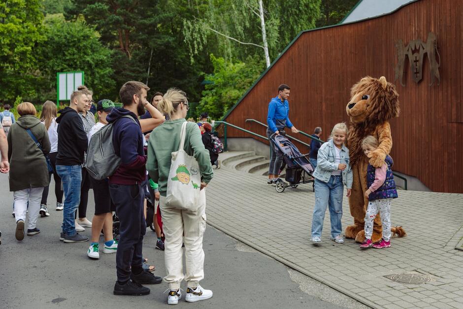 Po lewej grupa gości zoo w różnym wieku, po prawej dwie dziewczynki w wieku szkolnym pozują do zdjęcia z osoba przebraną w strój lwa