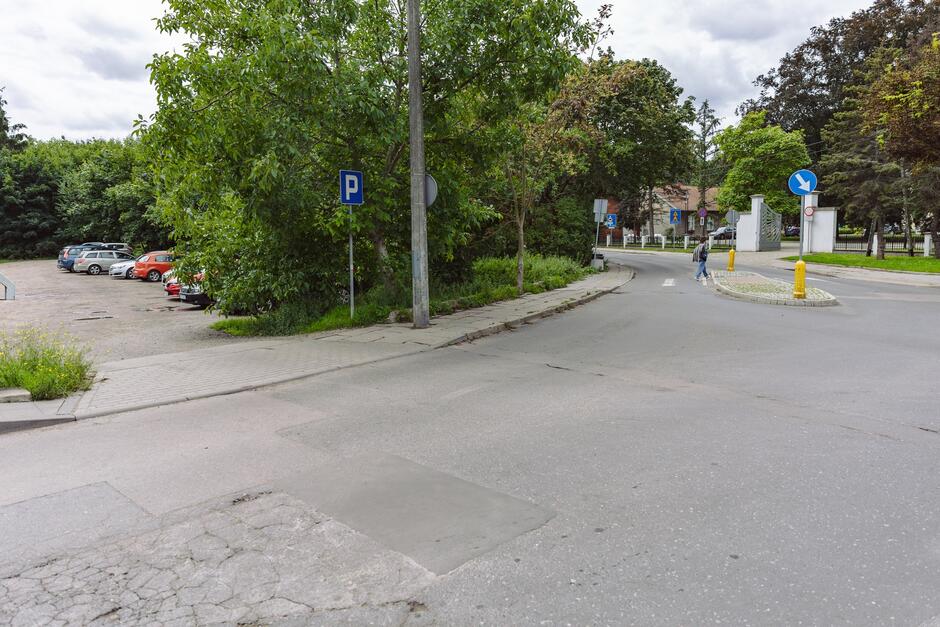 na zdjęciu fragment ulicy, po lewej w tle widać kilka zaparkowanych samochodów i zielone drzewa, po prawej w oddali widać przejście dla pieszych i dalej dużą bramę parkową