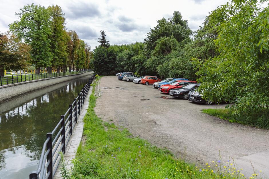 na zdjęciu widać gruntowy parking na którym jest zaparkowanych kilka samochodów, w tle widać zielone drzewa, po lewej fragment kanału rzeki radunia