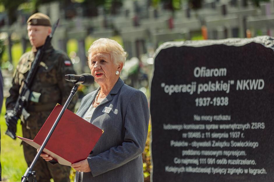 Starsza kobieta przemawiająca do mikrofonu przed pomnikiem