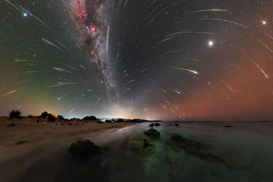 Zdjęcie nocne, w ciemnych kolorach, przedstawia w dolnej części krajobraz z niską roślinnością, w górnej - liczne świetliste kreski na niebie, zaznaczone przez spadające gwiazdy, i planety 