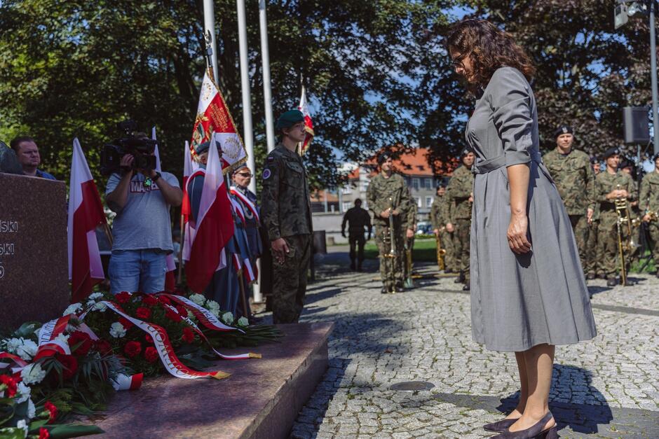 na zdjęciu prezydent gdańska w szarej eleganckiej sukience, kłania się przed pomnikiem, w tle stoją żołnierze