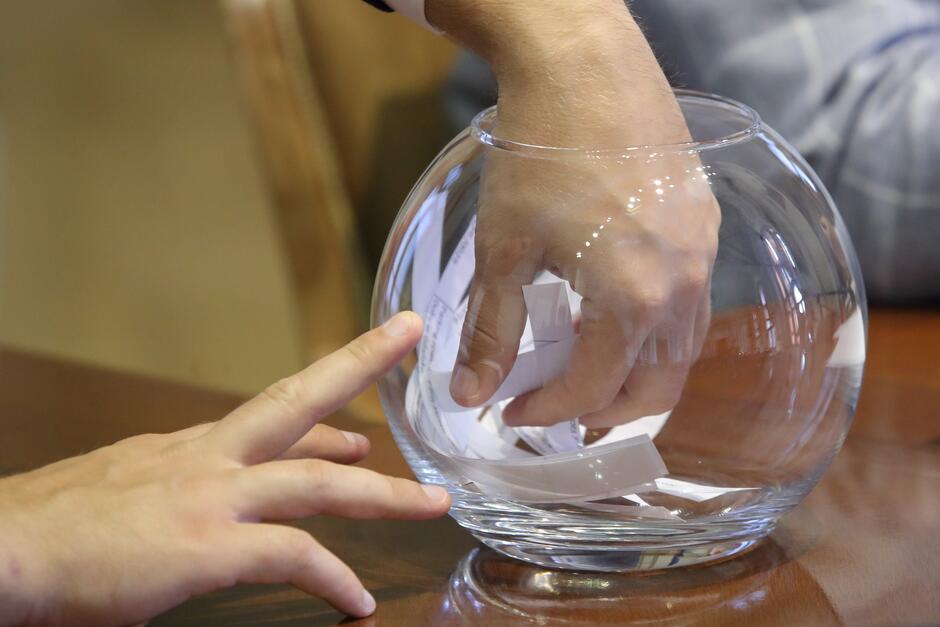 na zdjęciu ludzka ręka włożona do szklanego pojemnika okrągłego i głębokiego, w środku są krótkie białe karteczki