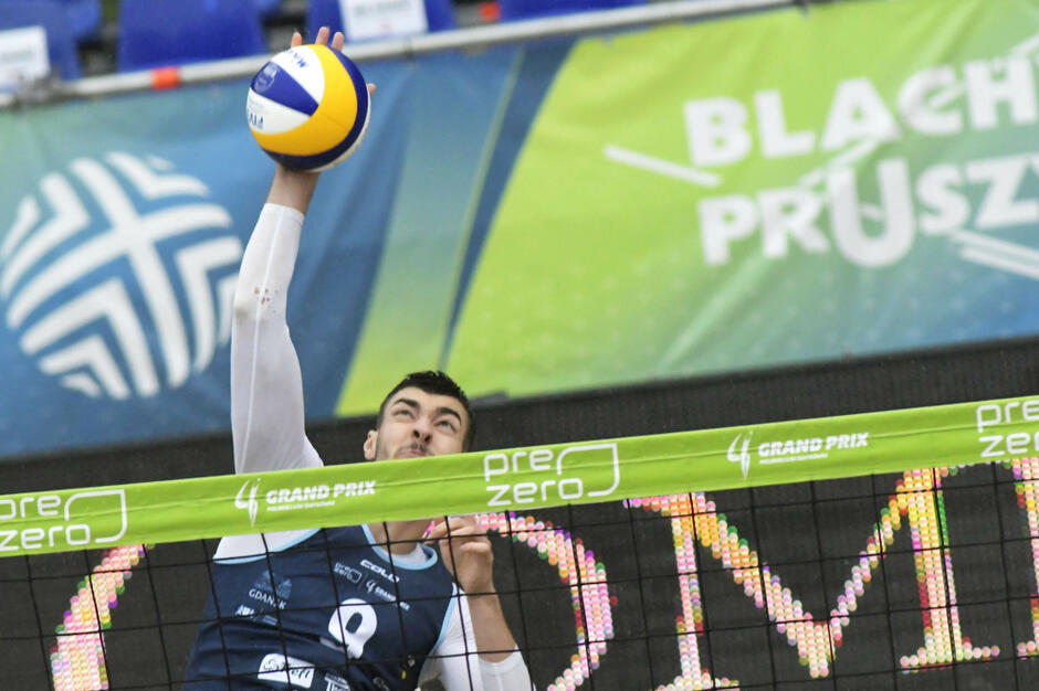 Siatkarz z ręką nad siatką, nad nim piłka. W tle reklamy