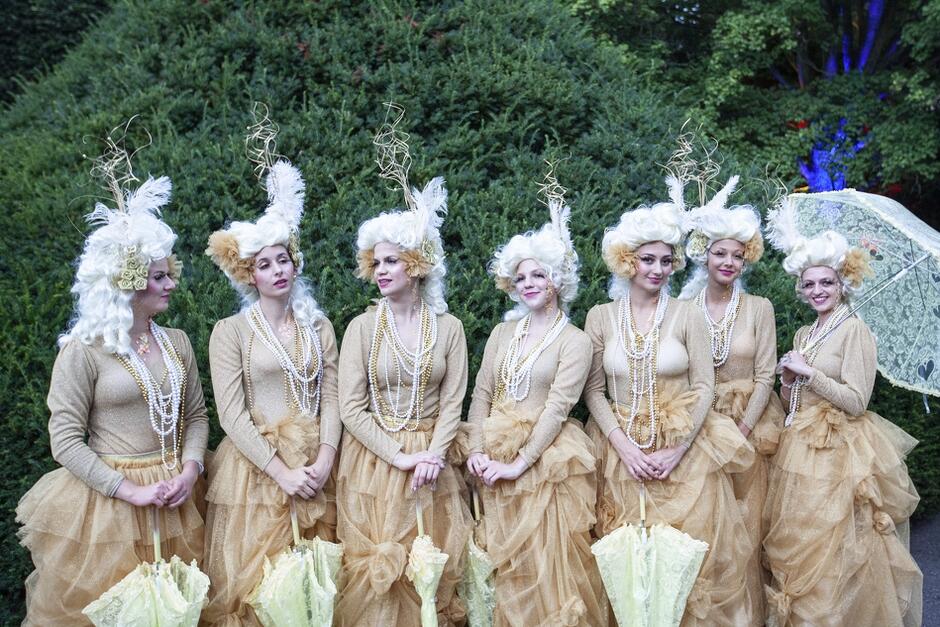 Siedem kobiet stoi pod zielonym parkanem, są ubrane w identyczne suknie z epoki Mozarta i białe wysokie peruki-koki. Trzymają w dłoniach parasolki chroniące przed słońcem