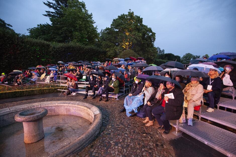 Zdjęcie wykonane w parku, na ławeczkach wokół fontanny, pod parasolami, bo pada deszcz, siedzą zasłuchani ludzie