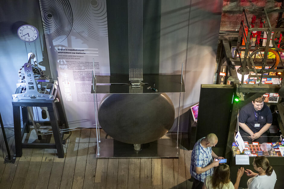 z boku widoczny stojący na podeście mechanizm zegara i wiszący nad nim cyferblat obok ogromnej średnicy wahadło podwieszone na kilku stalowych linach