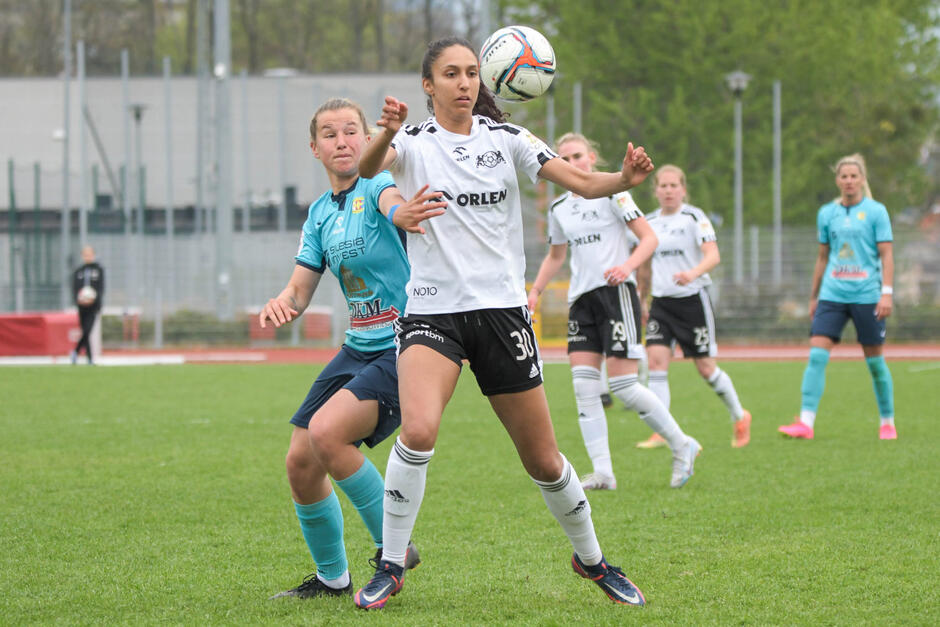 Dwie piłkarki walczą o piłkę na boisku (piłka na wysokości piersi jednej z nich). W tle inne zawodniczki
