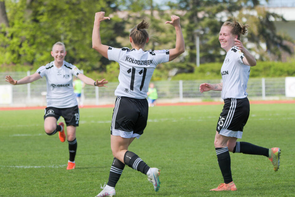 Piłkarka odwrócona plecami do obiektywu, ma podniesione ręce, kciukami wskazuje na plecy. Na koszulce nazwisko: Kołodziejek i numer 11. Dwie inne piłkarki dobiegają do niej z uśmiechami na twarzach