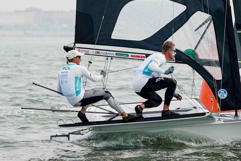 Dwóch żeglarzy na łodzi, są przykucnięci, trzymają linki w dłoniach 