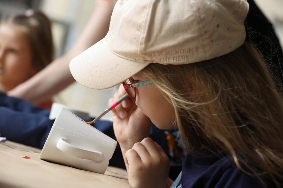 Dziewczynka w czapce z daszkiem maluje pędzelkiem wzorek na kubku porcelanowym