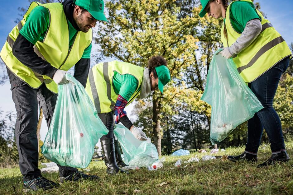 Trzy osoby w zielonych kamizelkach i czapkach z daszkiem są pochylone, w dłoni mają worki na śmieci, zbierają odpadki z trawnika 