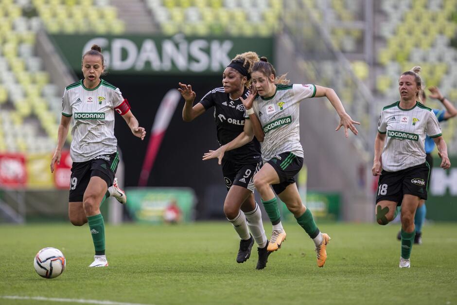 Dwie piłkarki walczą bark w bark o piłkę, po bokach dwie inne biegną. W tle trybuny