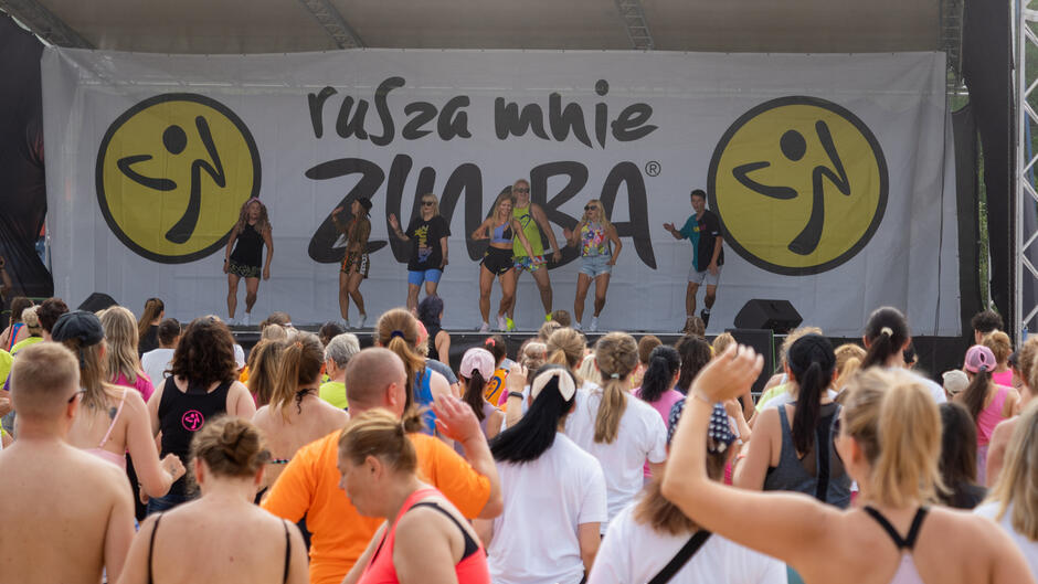 Kilkadziesiąt osób przed sceną. Na scenie kilka młodych kobiet pląsa, w tle baner z napisem RUSZA MNIE ZUMBA 