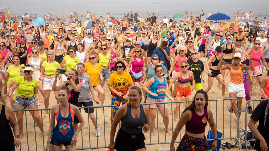 Na plaży kilkaset osób tańczy, na przedzie cztery młode kobiety w czarnych sukienkach, 