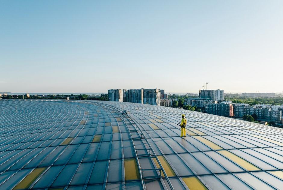 mężczyzna stoi na dachu stadionu, ubrany jest w kombinezon i kask, myje urządzeniem powierzchnię dachu 