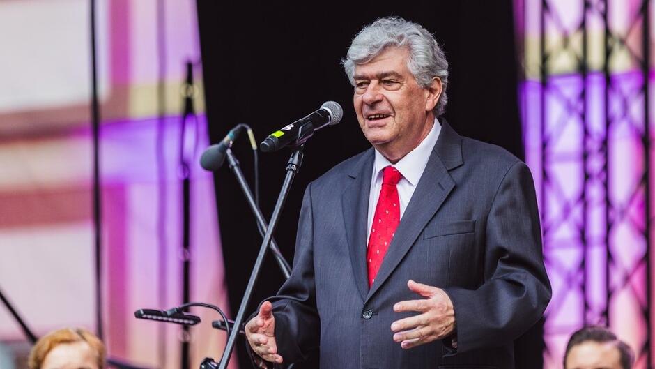 Starszy mężczyzna z gęstymi siwymi włosami, ubrany w garnitur i czerwony krawat przemawia na scenie stojąc do mikrofonu